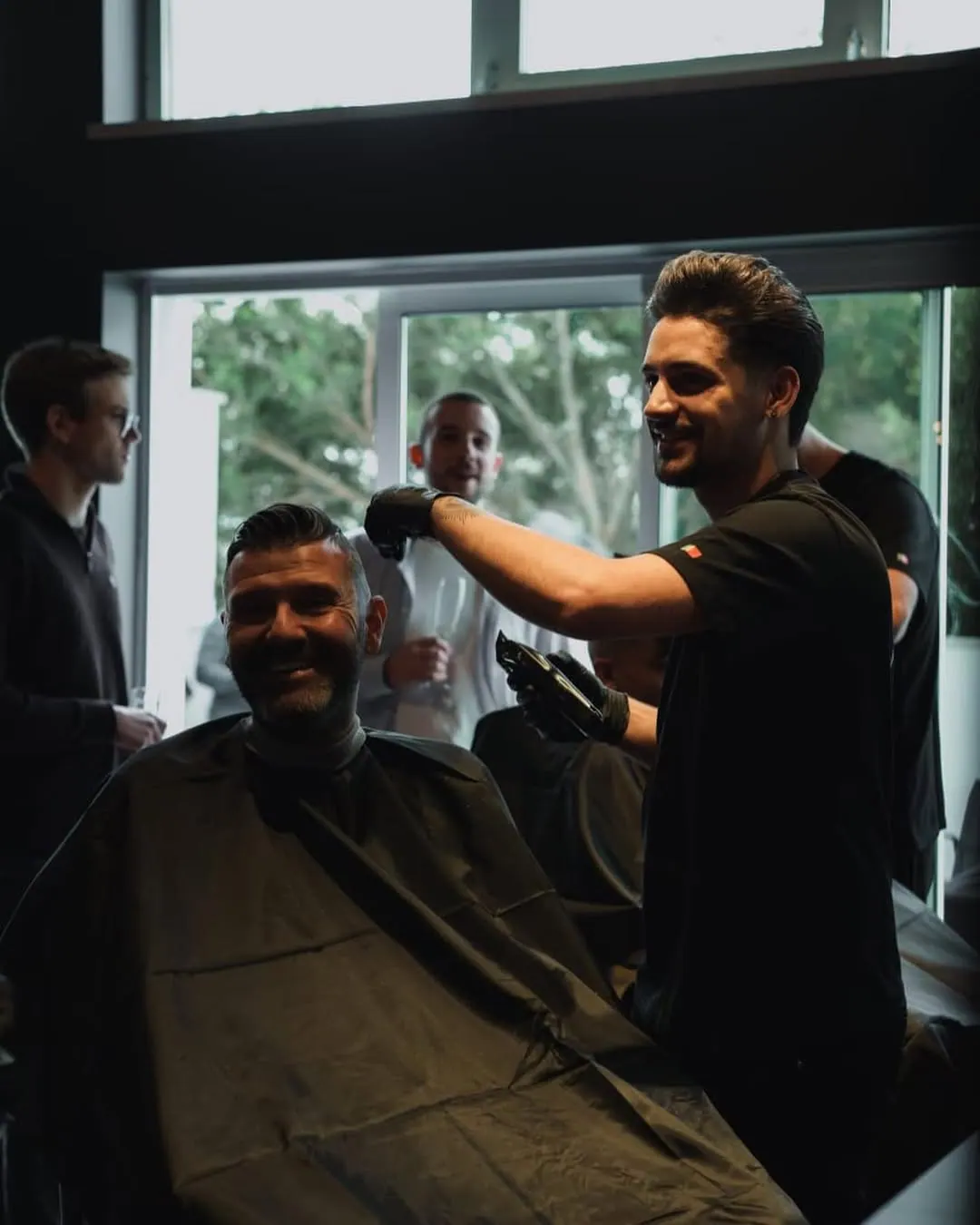 Barbeiro da Ganeno a sorrir enquanto corta o cabelo de um cliente, com amigos à janela